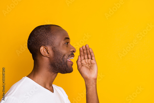 Closeup profile photo of cheerful dark skin guy good mood yelling empty space hold arm near mouth novelty information wear casual white t-shirt isolated bright vivid yellow color background photo