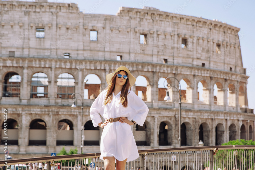 Beautiful young woman in colorful fashion dress alone in front of colosseum in Rome at sunset. Attractive girl tourist taking off elegant straw hat.
