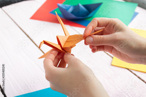 A girl puts together an origami crane and a boat made of colored paper. On a wooden background
