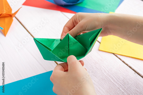A girl puts together an origami crane and a boat made of colored paper. On a wooden background