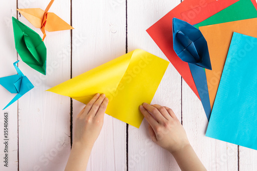 A girl puts together an origami crane and a boat made of colored paper. On a wooden background