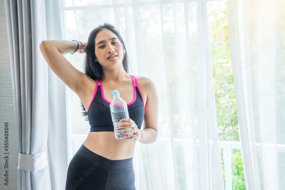 Asian woman wears a sports bra and holding a water bottle to rest after a morning workout in the house.Stay home and quarantine concept.