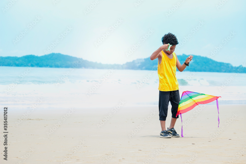 Cute kid having fun on sandy summer with blue sea, happy childhood boy playing colorful kite on tropical beach