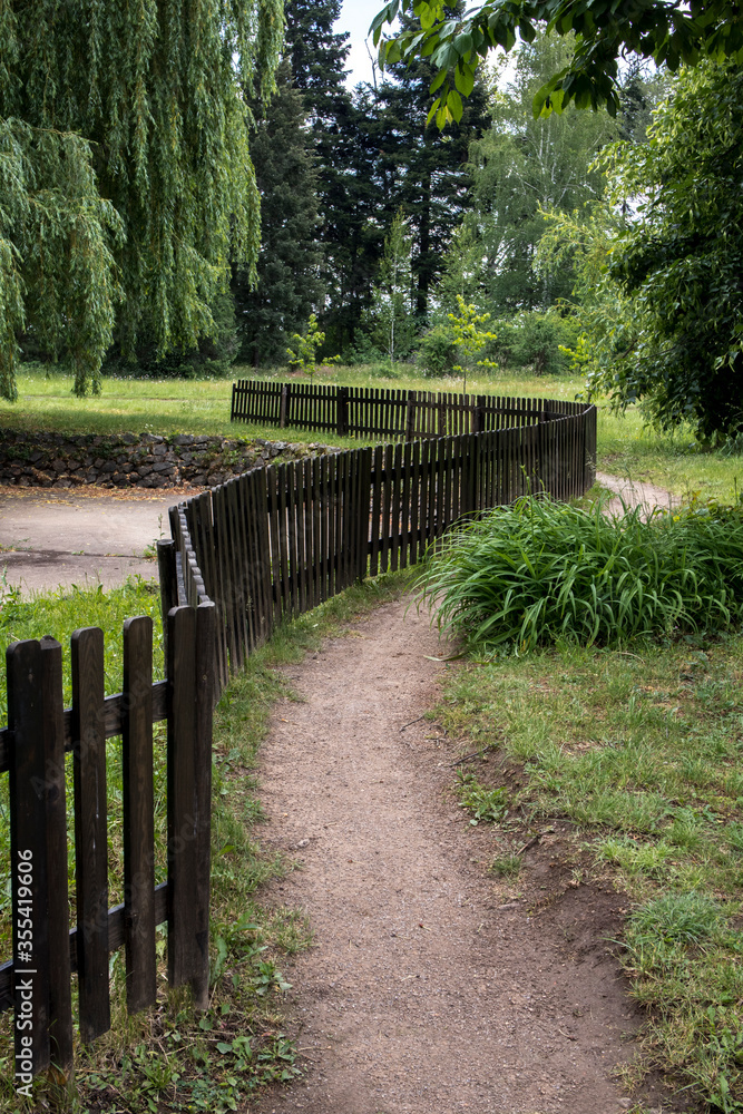 Curved path in the park with a black wooden fence, green landscape with tall trees.