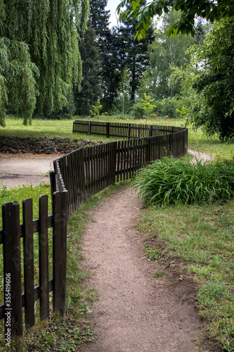 Curved path in the park with a black wooden fence  green landscape with tall trees.