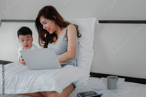 The mother works with her son in the bedroom at home. work at home, concept with a women sitting on the bed with laptop and holding a coffee mug.