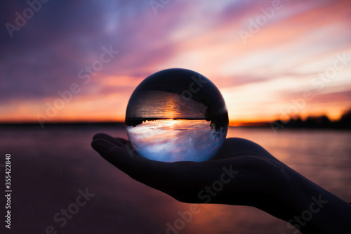 Beautiful scenic view through lens of crystal ball on water horizon at summer sunset. Clouds reflection in sphere. Palm of hand holding globe in outdoor.   photo