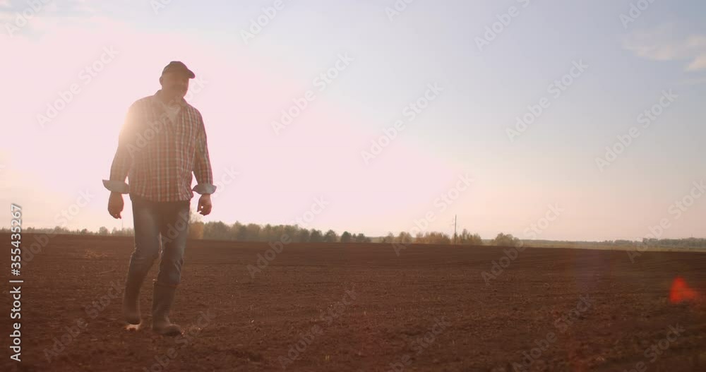 Slow-motion: senior male a tractor driver walks through a plowed field at sunset after work Slow-motion: senior male a tractor driver walks through a plowed field at sunset after work