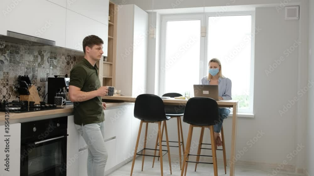 Young man is drinking a cup of tea while his wife is working at the computer, she is with a medicinal mask on face. Work from home and stay at home due the pandemic situation concept Young man is drinking a cup of tea while his wife is working at the computer, she is with a medicinal mask on face. Work from home and stay at home due the pandemic situation concept