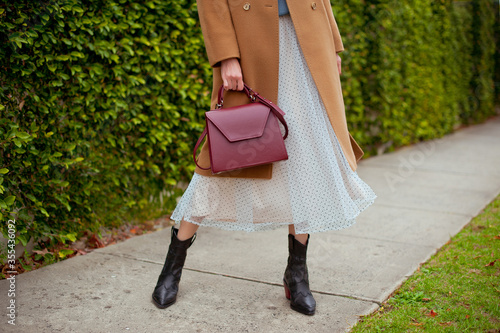 Detail of young fashionable woman wearing beige wool coat, tulle midi skirt and black high heel cowboy boots. She is holding stylish burgundy handbag in hands. Street style.