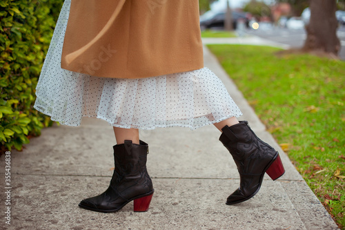 Detail of young fashionable woman wearing beige coat, tulle midi skirt and black high heel cowboy boots. Street style.