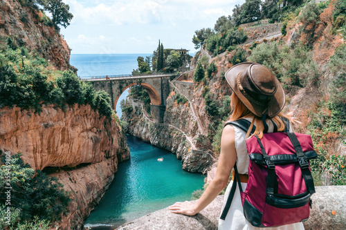 A girl in a cowboy hat with a backpack enjoys an incredibly beautiful view of a stone bridge over the gorge Fiordo di Furore. Little bay with paradise beach. Back view. Amalfi, Italy photo