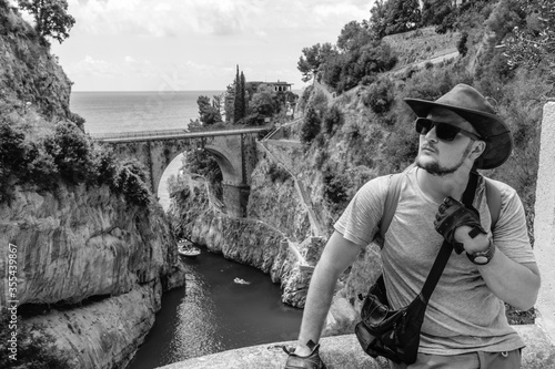 The handsome man traveler enjoys a wonderful view of the beach and the stone bridge. Sunglasses and a kangaroo leather cowboy hat. Travel and vacation. Fiordo di Furore, Amalfi, Salerno coast.
