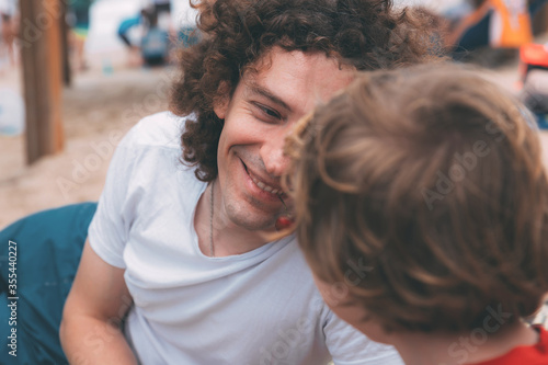 Father's day. Dad and son. Little son feeds dad ripe cherries on the beach. Happy family father and child. Young handsome man with long curly hair smiles and his son in the foreground from back