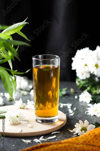 Yellow drink in a transparent tall glass on a table with flowers. The glass stands on a wooden stand on a dark table next to white flowers