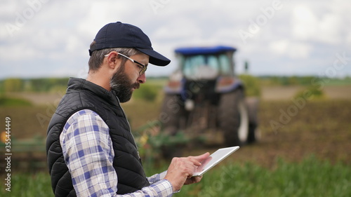 Farmer uses a specialized app on a digital tablet PC on the background of working tractor with a cultivator in the field