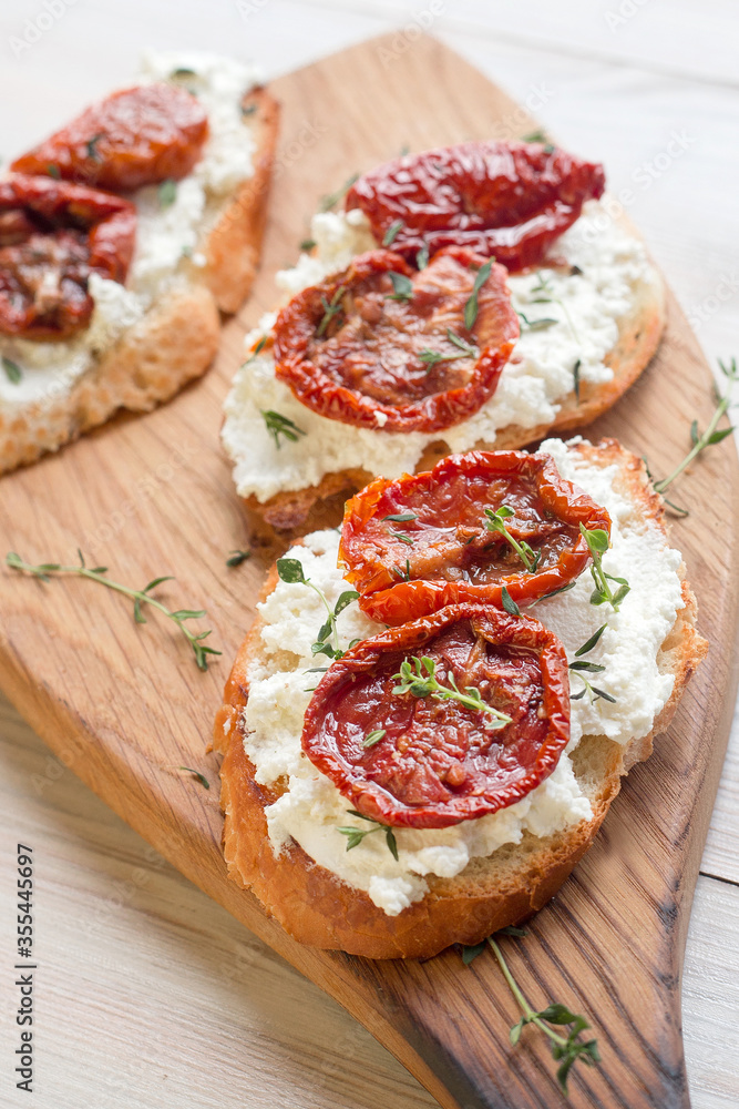 Toast (italian bruschetta) with sun-dried tomatoes and soft curd cheese with fresh herbs on wooden board, light background, closeup view, soft selective focus
