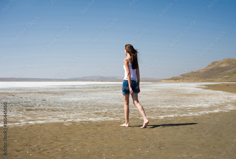 A young girl goes barefoot along the shore of a salt lake. Back view