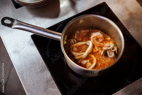 Sea food soup with shrimps, prawns, squids, little octopus is boiling, cooking in the saucepan on the induction cooker in the kitchen. Tom Yam or Bouillabaisse soup.