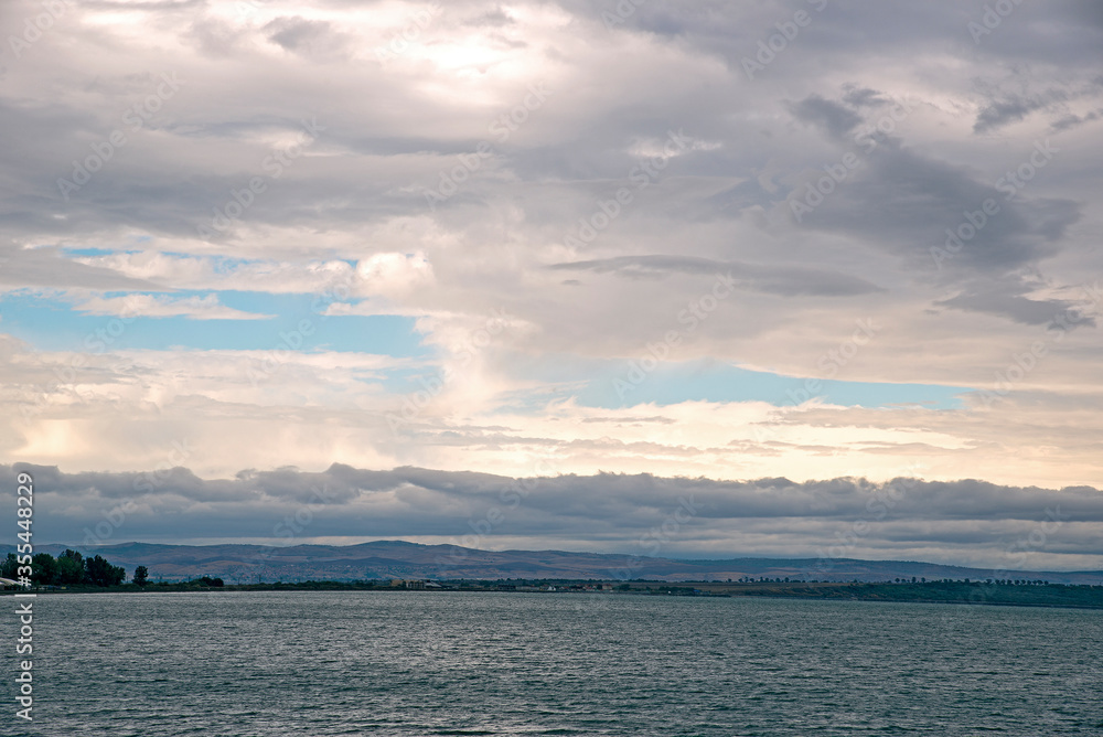 Gray rain clouds hang low over the calm surface of the sea. Forest and hills in the distance. Gray rain clouds hang low over the calm surface of the sea. Forest and hills in the distance.