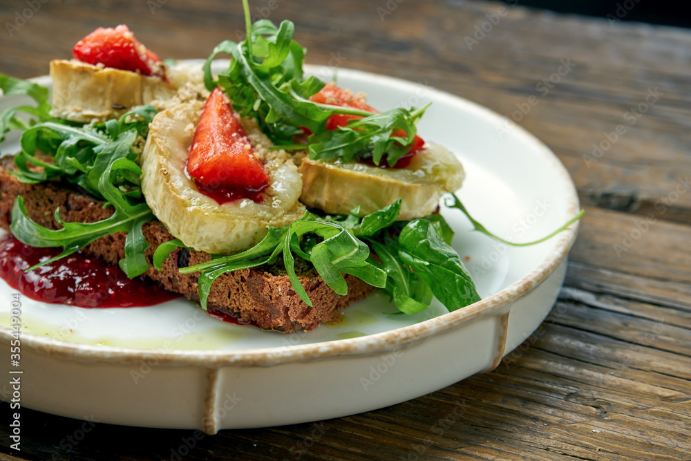 Appetizing toast with goat cheese, red jam, arugula and strawberries with rye bread on a plate on a wooden background
