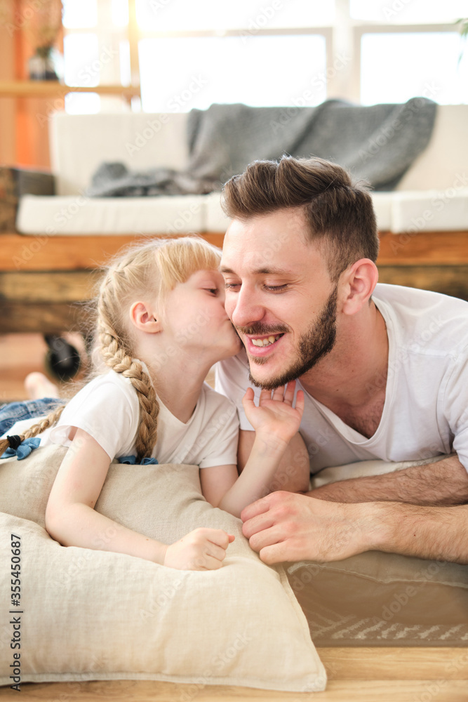 Father and daughter. Handsome young man and little cute girl lie on the carpet at home, hug, kiss, have fun. Dad and child laugh. Father's day.