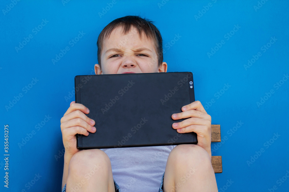 The boy is sitting on a chair and holding a tablet, looking at the tablet on a blue isolated background The boy is sitting on a chair and holding a tablet, looking at the tablet on a blue isolated background