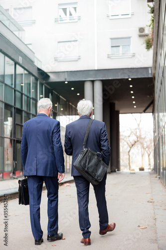 Two senior gray haired Caucasian businessmen in dark blue suits walking away from the camera in front of an office building. Rear view