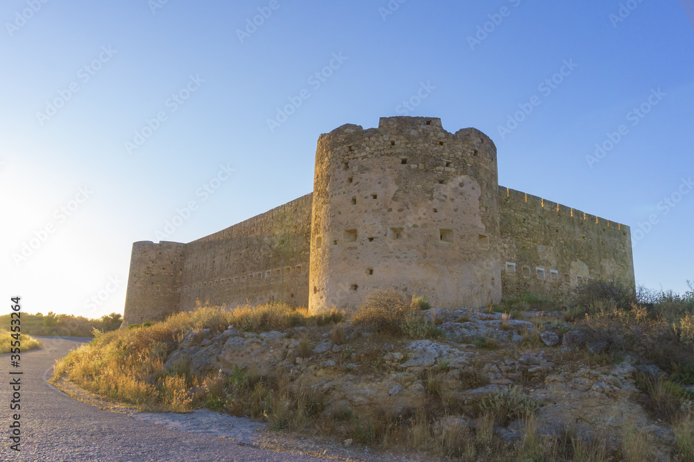 Impressive medieval castle with fortified towers and driveway on hill at sunset time, against clear blue sky.