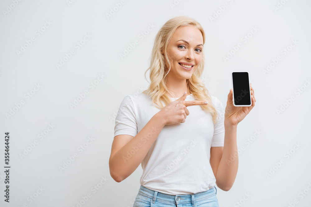 happy woman pointing at smartphone with blank screen while looking at camera isolated on white happy woman pointing at smartphone with blank screen while looking at camera isolated on white