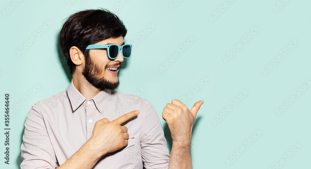 Studio portrait of young smiling guy wearing shades, pointing with fingers on empty background of aqua menthe color with copy space. Studio portrait of young smiling guy wearing shades, pointing with fingers on empty background of aqua menthe color with copy space.