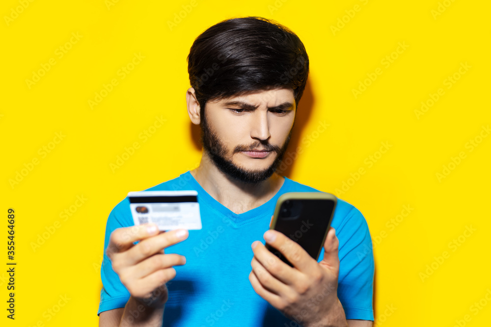 Studio portrait of young thoughtful guy using smartphone and credit card on yellow background.