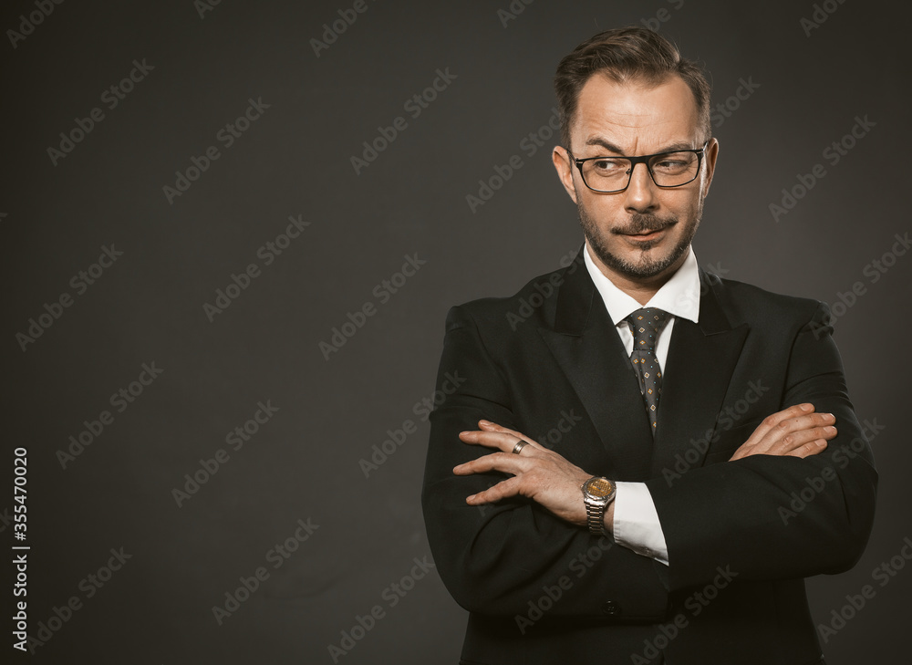 Confident businessman cunningly looks away with his arms crossed. Caucasian middle-aged man standing against a gray wall with copyspace or space for text on the left. Toned image. Confident businessman cunningly looks away with his arms crossed. Caucasian middle-aged man standing against a gray wall with copyspace or space for text on the left. Toned image.