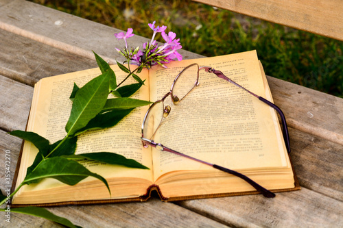 An open book lies on a wooden bench in the open air, with a purple flower and glasses.