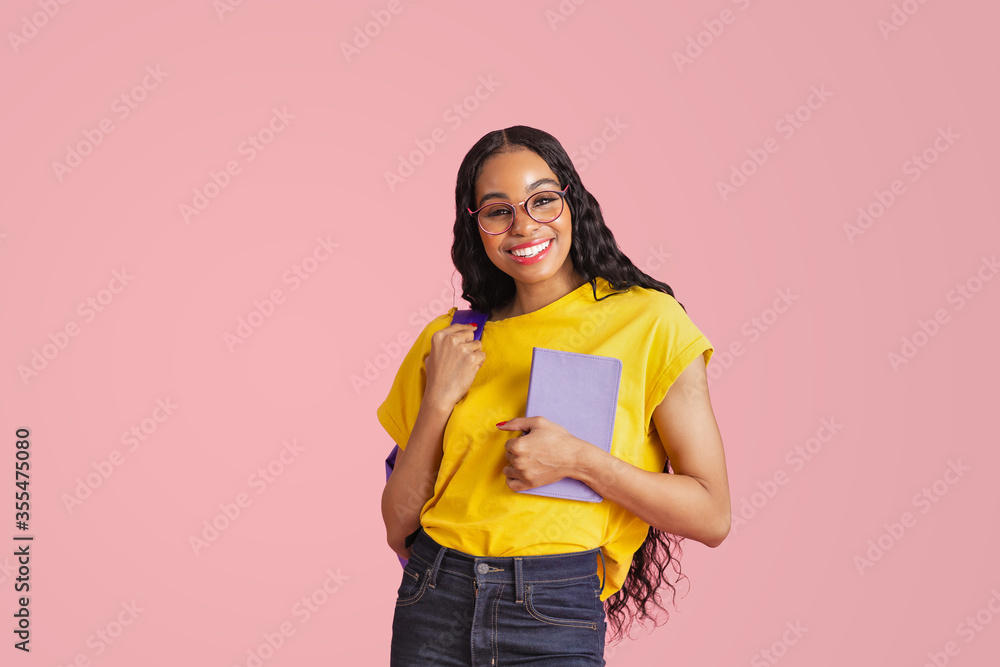 Portrait of a friendly young female student with book, backpack and glasses, isolated in pink studio copy space Portrait of a friendly young female student with book, backpack and glasses, isolated in pink studio copy space