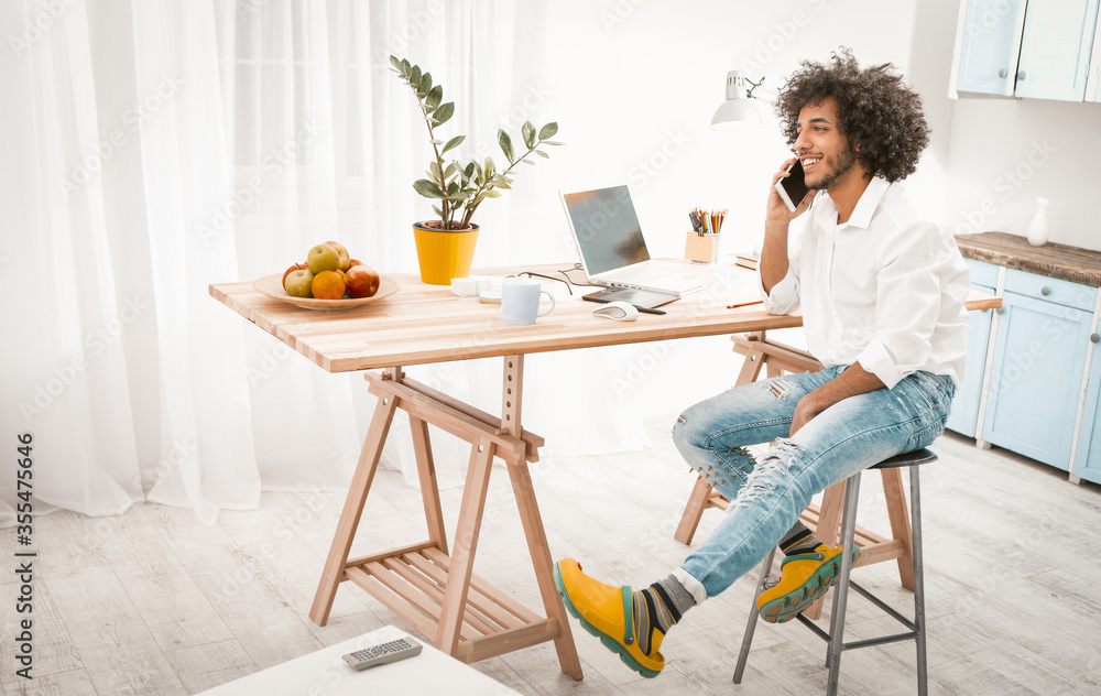 Creative man talks on mobile phone working from home. Young Arab guy in casual works in studio home kitchen interior. Toned image. Creative man talks on mobile phone working from home. Young Arab guy in casual works in studio home kitchen interior. Toned image.