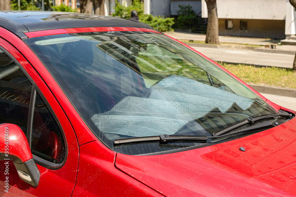 Foil sun shades covered a panel of a red car parked on the street on a sunny summer day. Reflective sun shield made of metallic silver foil protects vehicles from direct sunlight.