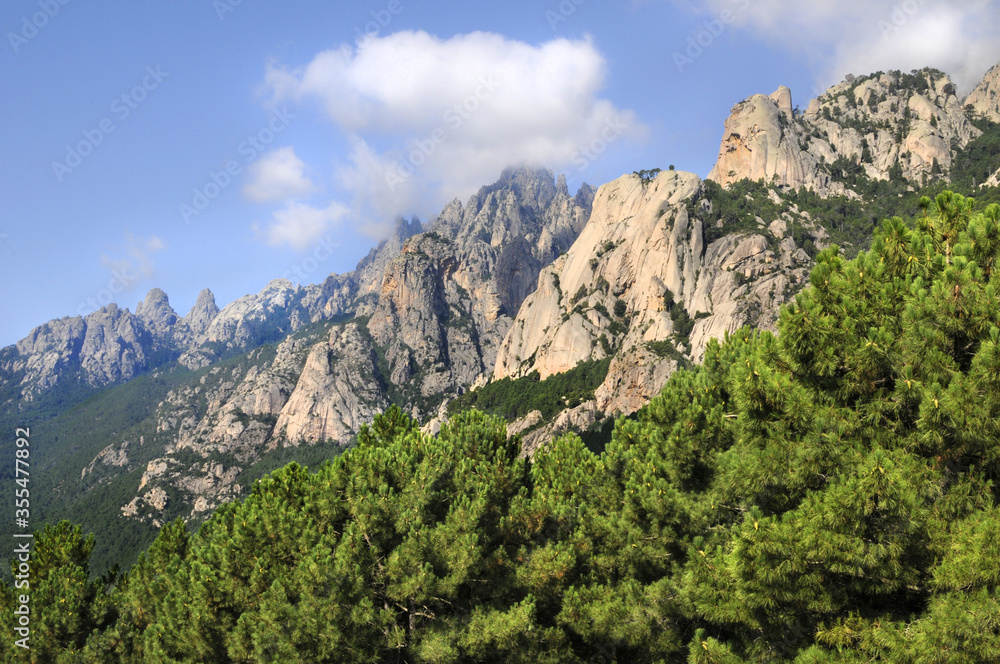 Pins Laricio et Aiguilles de bavella en Corse, sur fond de ciel bleu nuageux et nature sauvage et abrupte des rochers formant une montagne découpée au profil accidenté et dentelé.