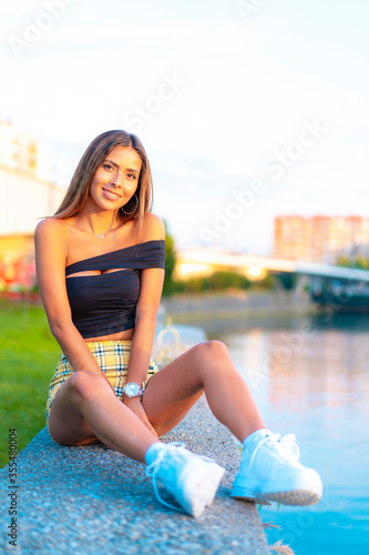 Lifestyle, a young brunette Caucasian woman in a tight black top and yellow skirt smiling, sitting on the city river at sunset