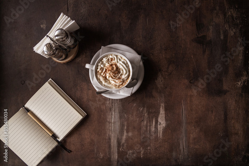 Dark photo. Cappuccino with whipped cream and cinnamon next to a notebook. Cozy coffee shop, flat, selective focus
