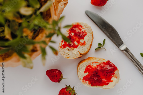Slices of cut baguette bread with butter and strawberry jam, strawberries, metal butter knife and green thyme plant leaves on a pastel white background in a flat lay composition