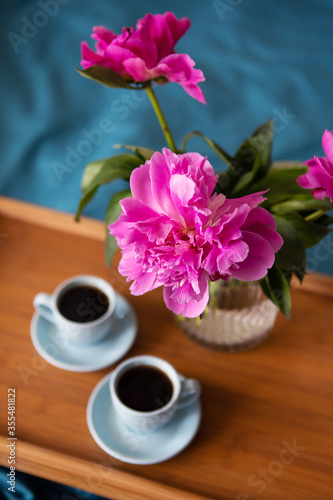 Fototapeta Naklejka Na Ścianę i Meble -  Beautiful pink peonies in a glass vase and two cups of coffee are standing on a wooden tray in the bed. Close-up.
