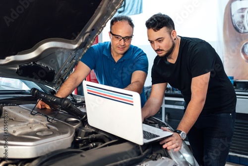 The mechanic man points to his laptop computer which shows the fault of the car. The two men look at the computer for the diagnosis. Diagnostic tool of failure