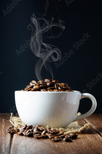 Coffee beans in a white cup over dark background and on a wooden surface