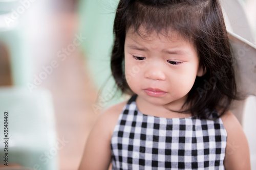 Little girl made a sad face from regret. Asian children bend their heads to cry. There are tears in eyelashes. Child frowns from uneasiness. Kid is wearing black and white checkered shirt. 3 years old