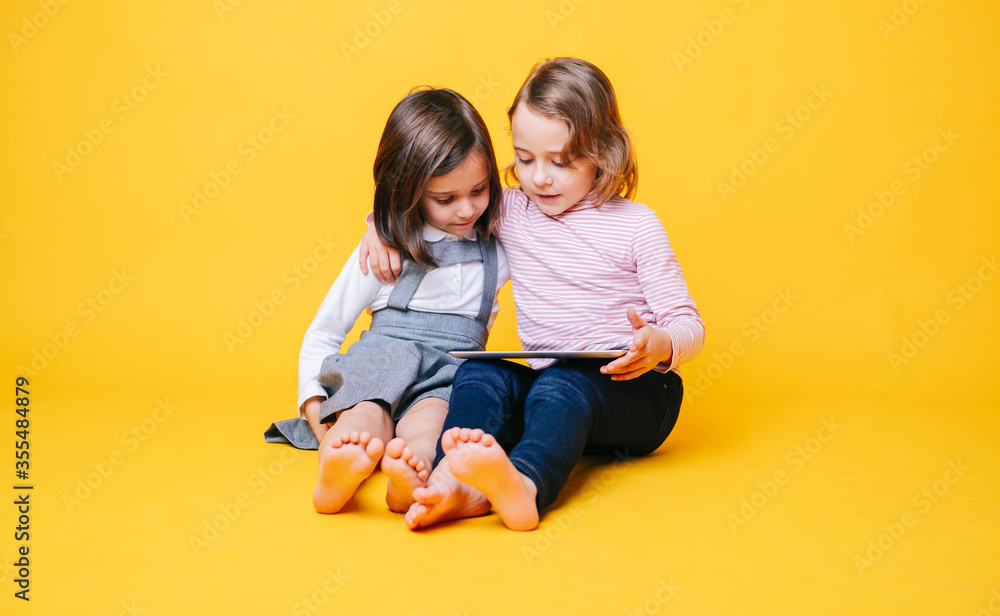 Two girls use a digital tablet on an isolated yellow background with copy space Two girls use a digital tablet on an isolated yellow background with copy space