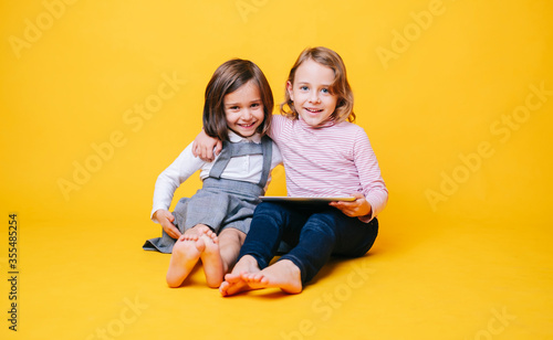 Two girls use a digital tablet on an isolated yellow background with copy space