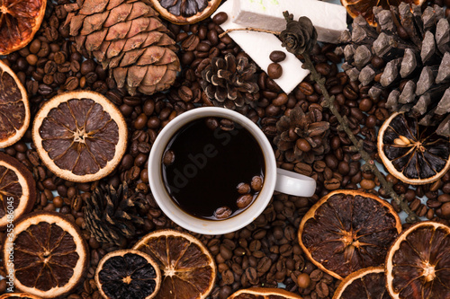A white Cup of coffee on coffee beans, on a light blue wooden table. Wooden table. Fir cones on the table. white marshmallow. Dried citrus. Brown background.