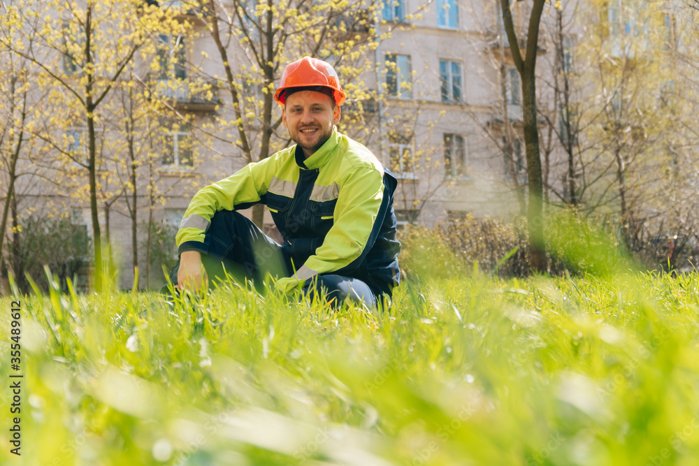 European man smiles in special clothes. The builder is sitting on the grass. Rest break from work European man smiles in special clothes. The builder is sitting on the grass. Rest break from work