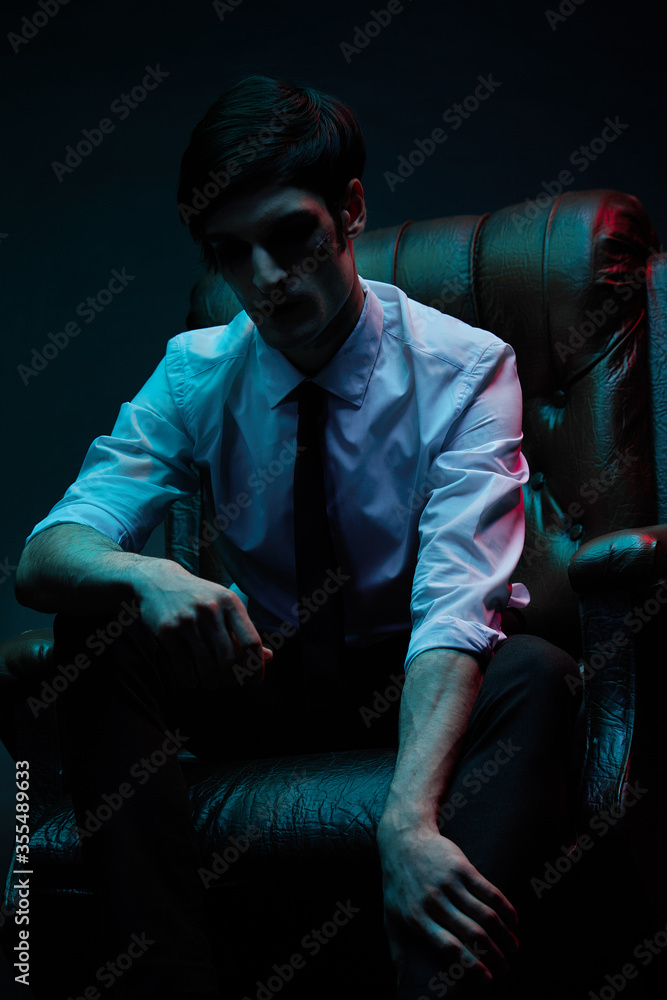 Portrait of a man in blue and red contrasting lighting in a white shirt with a tie and a facial injury, posing sitting in a leather chair. Futuristic neon lighting Portrait of a man in blue and red contrasting lighting in a white shirt with a tie and a facial injury, posing sitting in a leather chair. Futuristic neon lighting